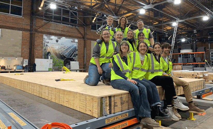 solar-decathlon-students Photo of 11 students, wearing safety glasses and vests, in a warehouse with construction materials.