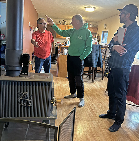 Three people stand around a wood-burning stove in a house.