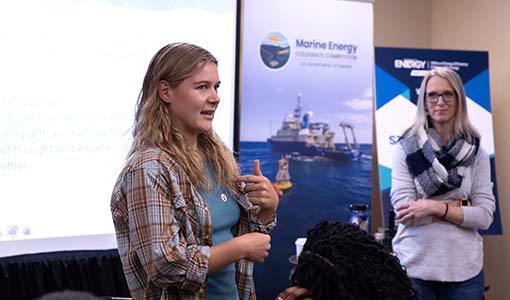 A person speaks in front of posters and a presentation screen as another person watches.