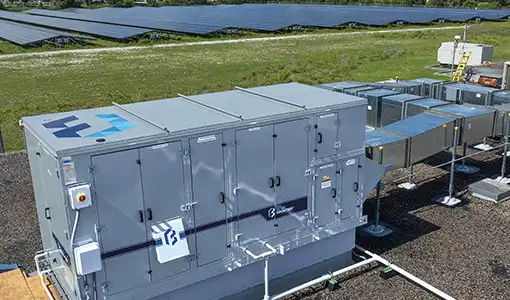 Photo of a large metal air conditioning unit with vents and access panels. The unit is adjacent to a field of photovoltaic panels.